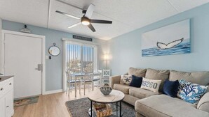 A living room with a ceiling fan, a dining area with a table and chairs, a sofa with decorative pillows, and a wall with a whale tail painting.