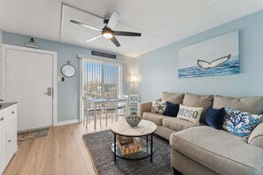 A living room with a ceiling fan, a dining area with a table and chairs, a sofa with decorative pillows, and a wall with a whale tail painting.
