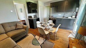 Una sala de estar moderna con sofá, mesa de comedor para cuatro personas y una cocina con armarios oscuros y salpicadero de azulejos.