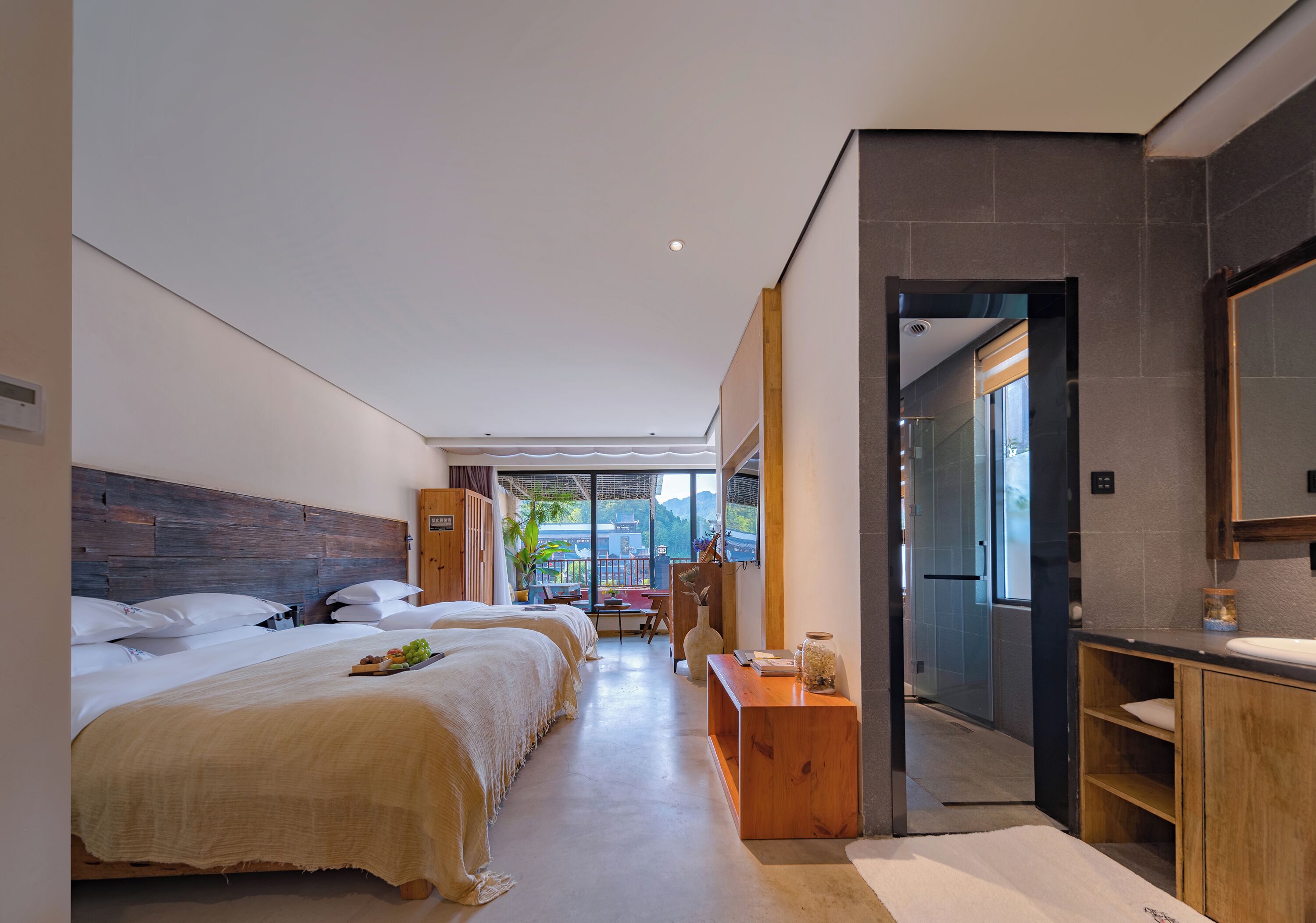 A modern hotel room with a large bed, a wooden headboard, a small desk with a vase, and a view of the outdoors.
