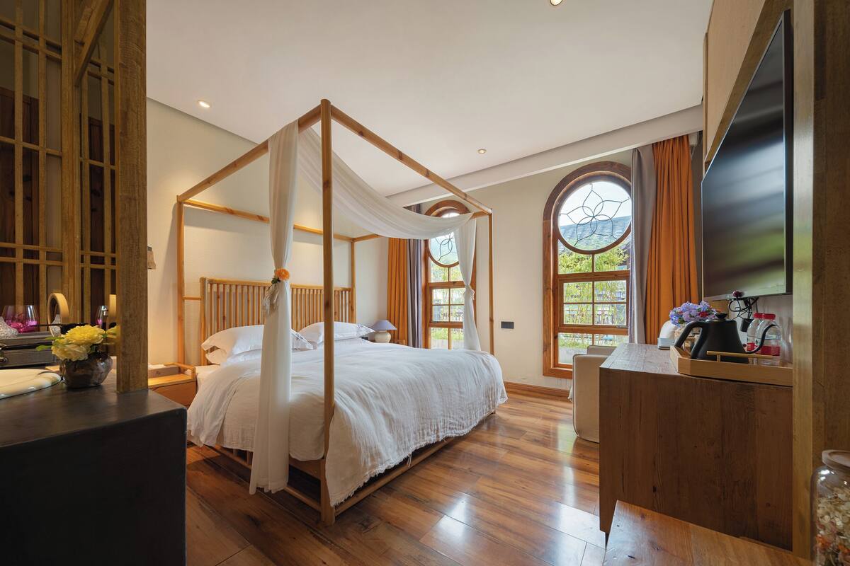 Ein Schlafzimmer mit Himmelbett, Holzboden, Schreibtisch mit Wasserkocher und großem Fenster mit Vorhängen.
