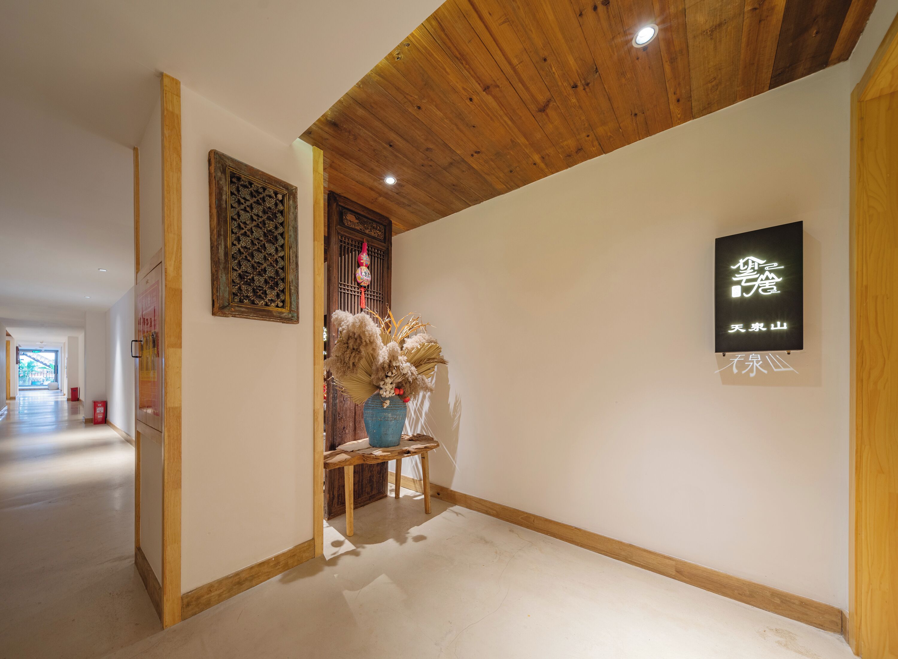 A hallway with wooden ceiling beams, a decorative wall piece, a blue vase with dried plants, and a sign with Chinese characters.