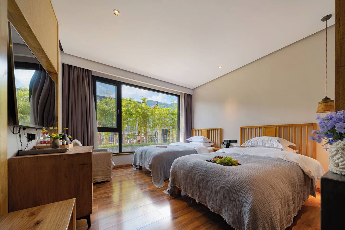 Ein modernes Hotelzimmer mit zwei Betten, einem Holztisch, einem großen Fenster mit Blick ins Grüne und einem an der Wand befestigten Flachbildfernseher.