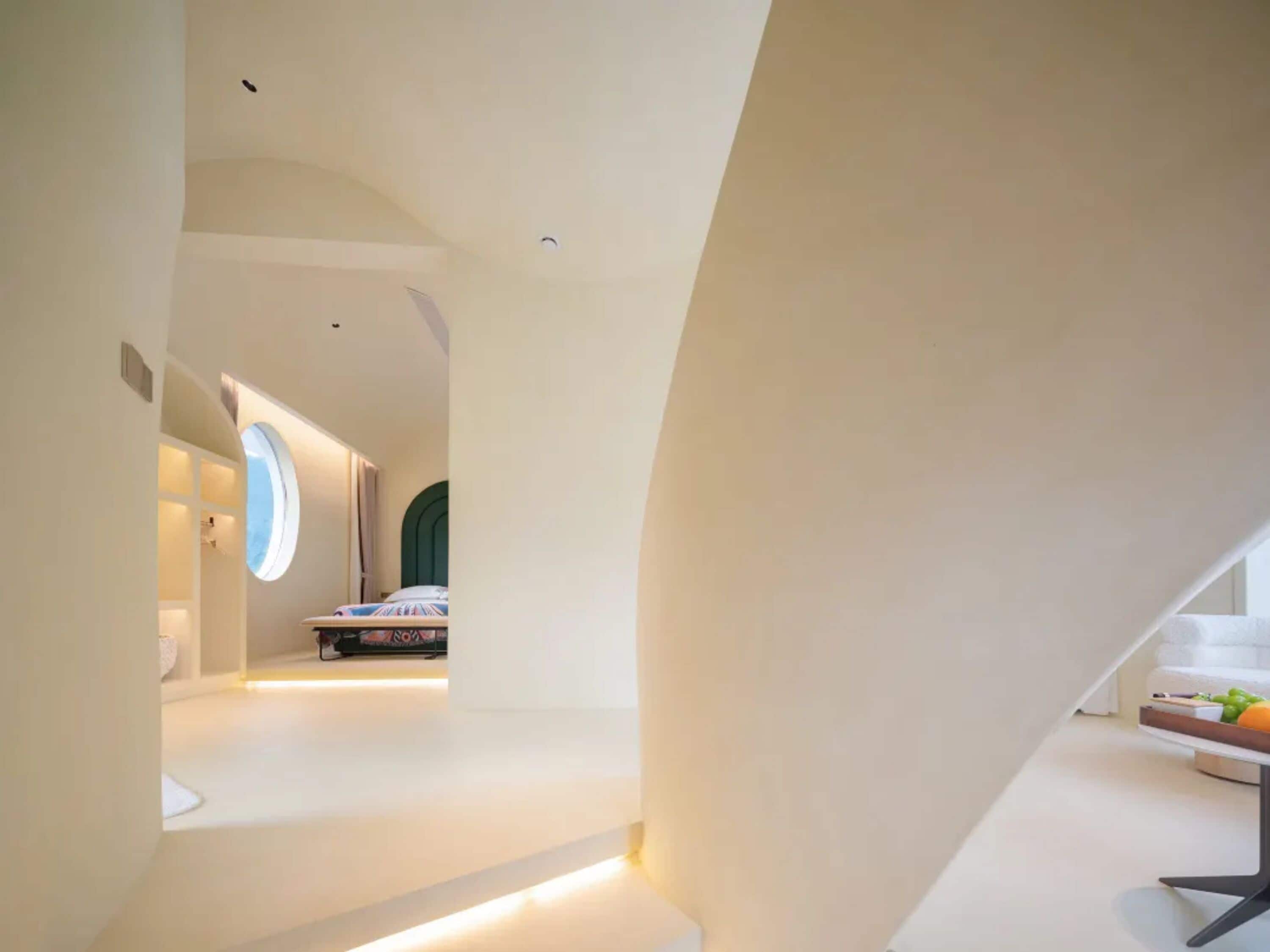 A modern interior with a curved white wall, a bed with a green headboard, and a small table with fruits.