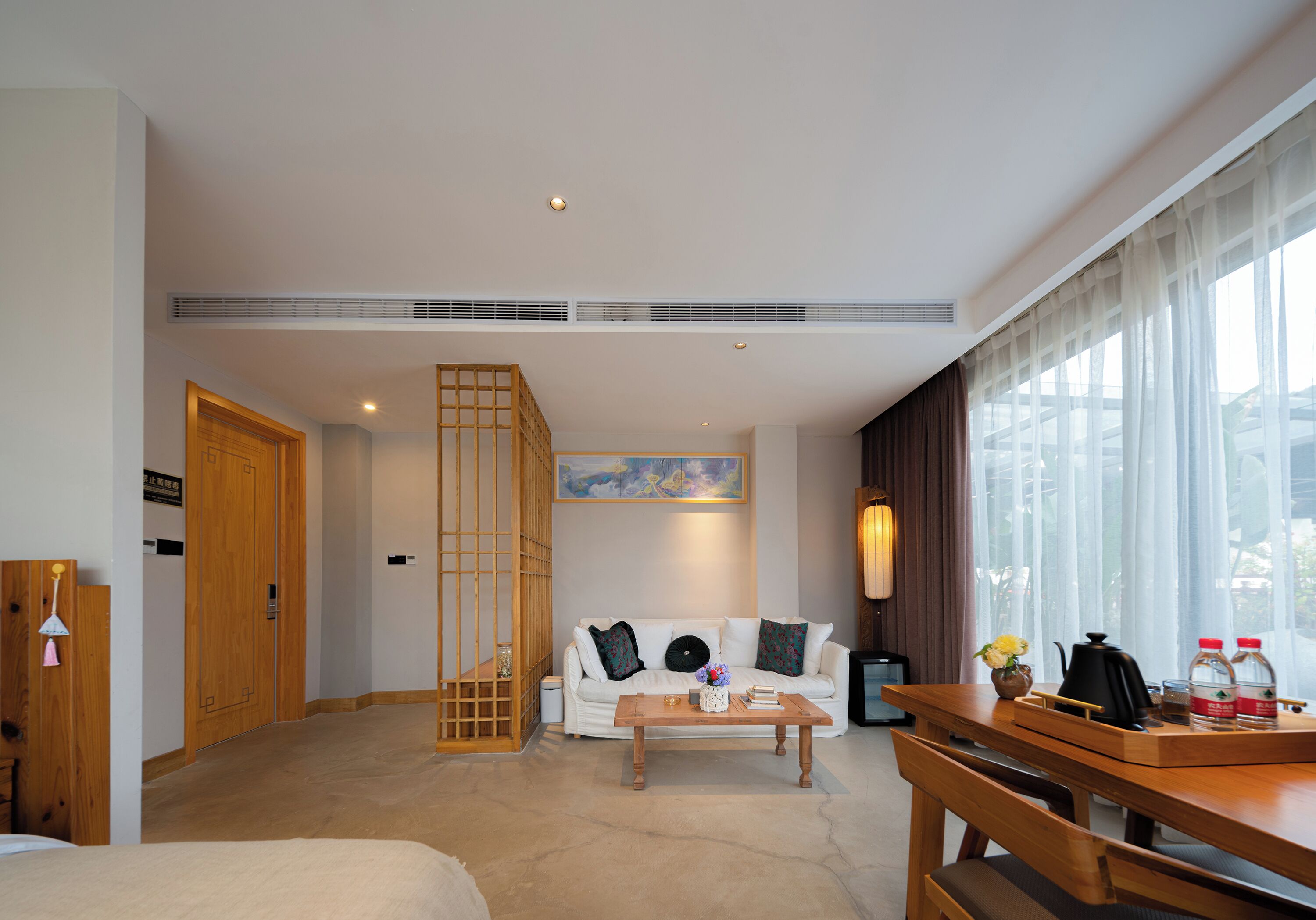 Une chambre d’hôtel avec un canapé, une table basse et un coin repas comprenant une table et des chaises. Il y a une fenêtre avec des rideaux, une lampe et un tableau au mur.