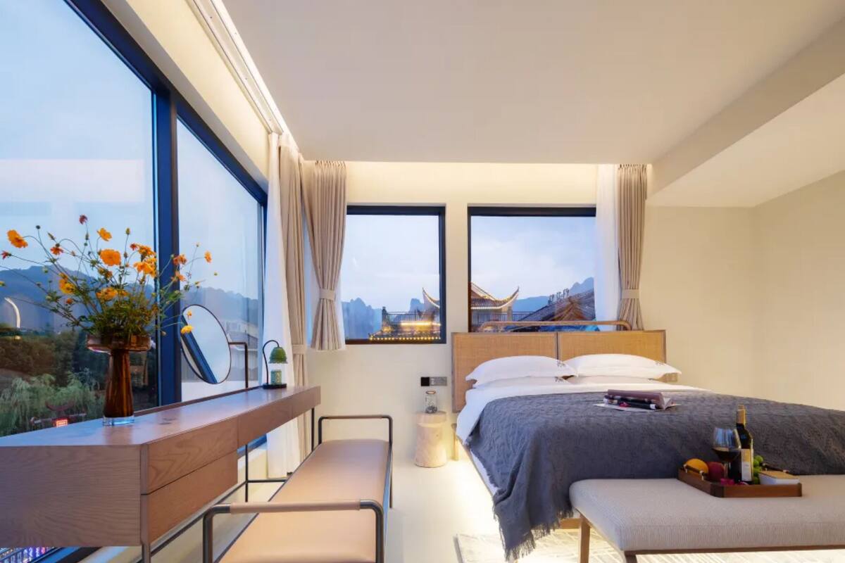 Ein modernes Hotelzimmer mit einem großen Bett, einem Schreibtisch mit Stuhl, einem kleinen Tisch mit einer Flasche und Obst sowie großen Fenstern mit Blick auf Berge und traditionelle Architektur.
