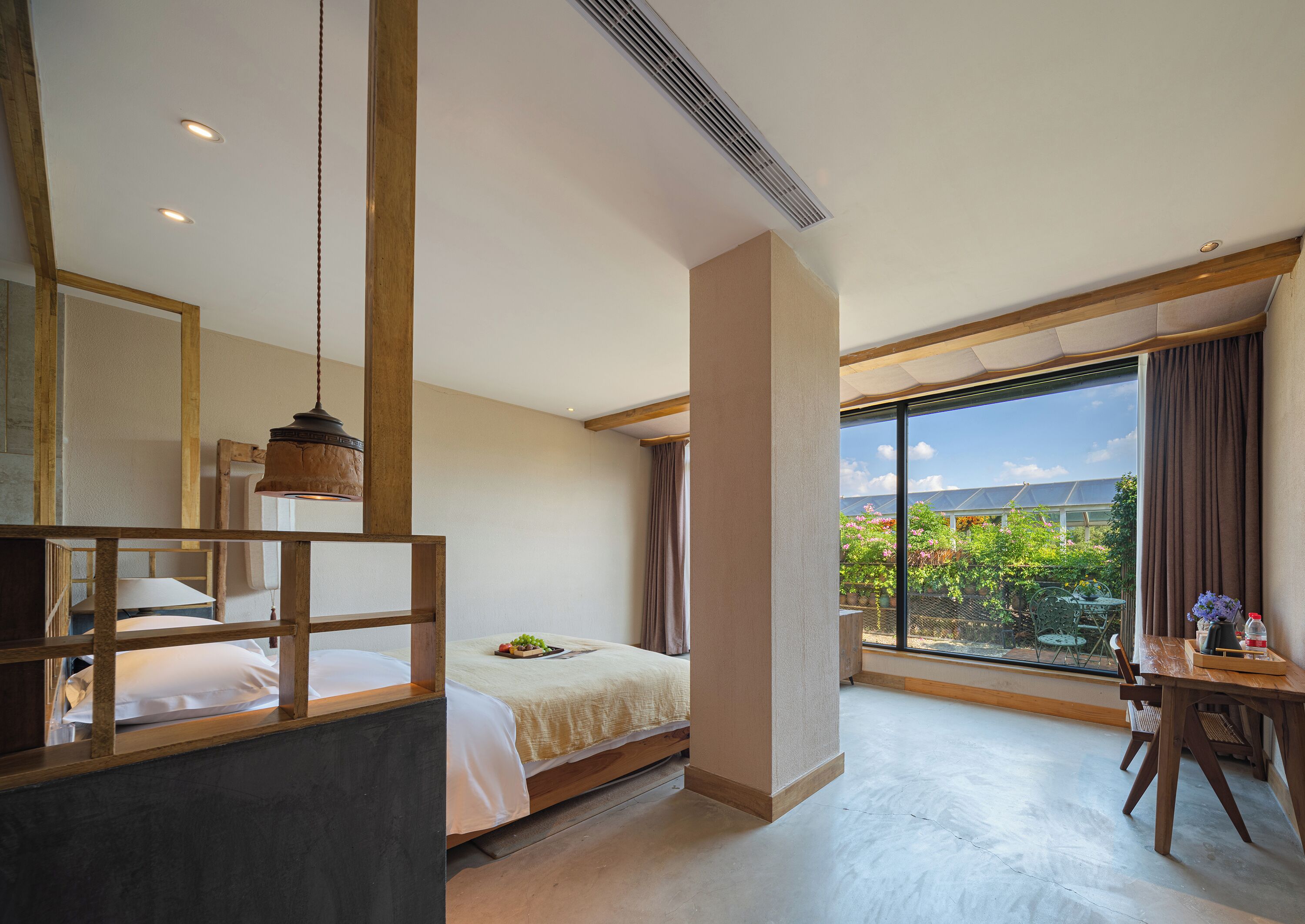 A bedroom with a bed, a desk, a chair, a large window with a view of greenery, and a ceiling vent.
