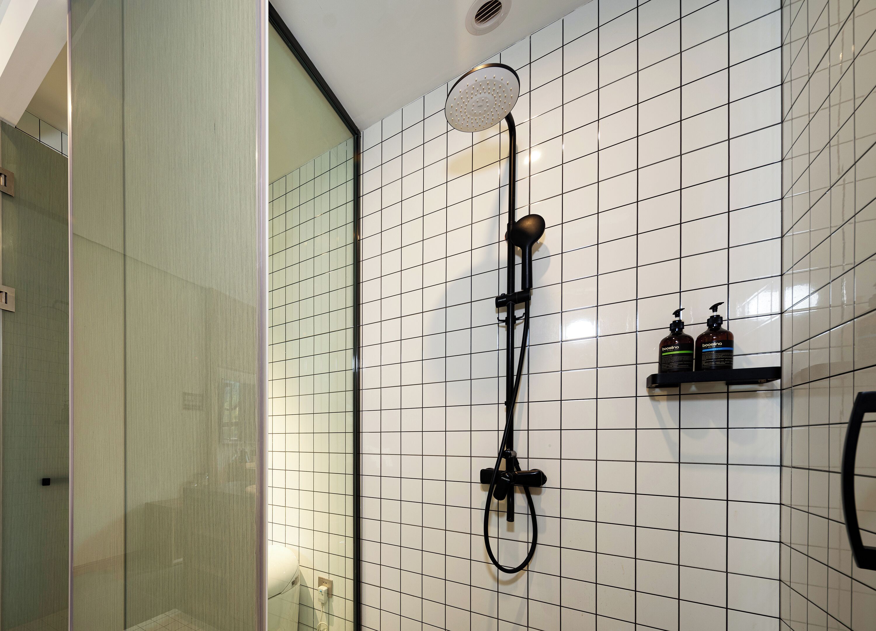 A bathroom with white tiled walls, a glass shower enclosure, a black showerhead, and a shelf with two bottles of lotion.