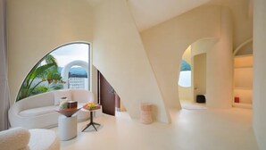 Un intérieur moderne avec un plafond incurvé, un canapé blanc, une petite table sur laquelle sont disposés des fruits, et une vue sur un immeuble à travers une grande fenêtre.