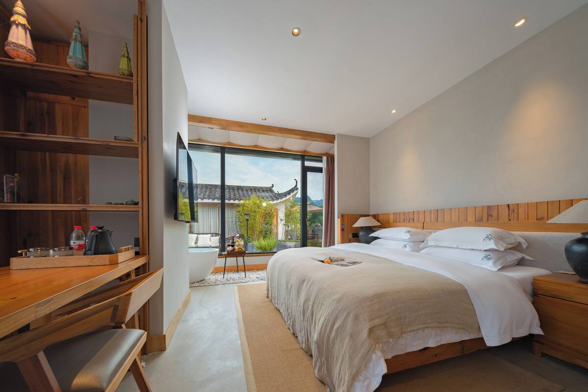 Ein modernes Schlafzimmer mit einem großen Bett, einem hölzernen Kopfteil und einem Schreibtisch mit Stuhl. Ein Fenster bietet Blick auf ein traditionelles Gebäude.