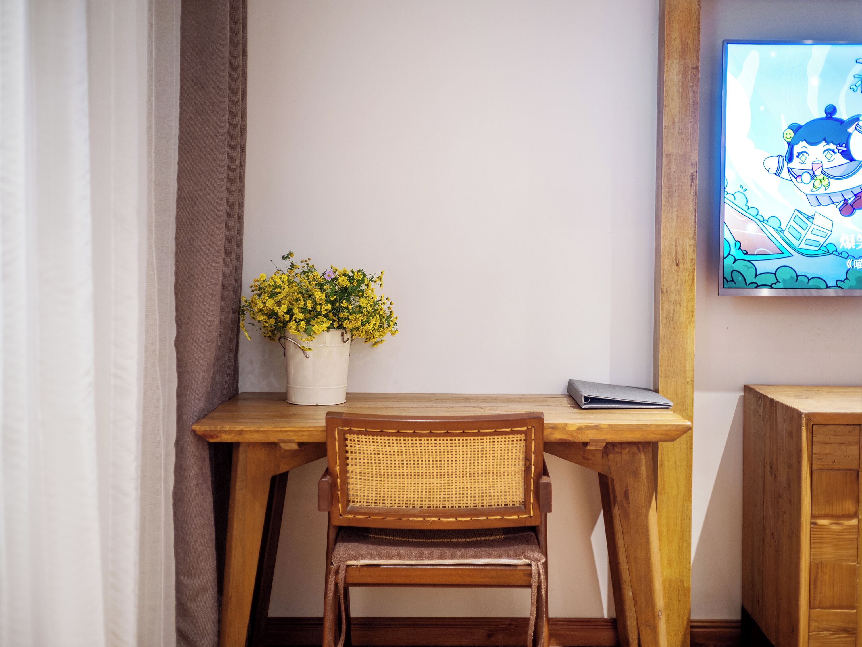 A wooden desk with a wicker chair, a white vase with yellow flowers, and a television on the wall.