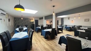 Ein Restaurant mit mehreren Tischen, jeder Tisch mit einer blauen Tischdecke und weißen Unterlagen. Schwarze Stühle stehen um die Tische, und Pendelleuchten hängen darüber. Der Boden ist Holzboden, die Wände sind grau und weiß gestrichen.