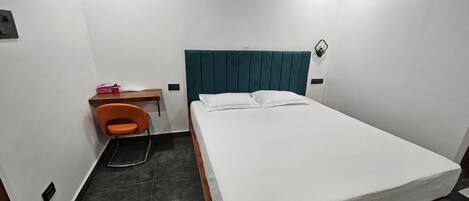 A small, single-bed room with a teal headboard, a wooden desk, an orange chair, and a white bedspread.