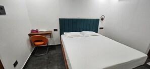 Una habitación pequeña con una cama individual, cabecera color turquesa, escritorio de madera, una silla naranja y colcha blanca.