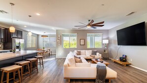 A spacious living area with a large sofa, wooden bar stools, a kitchen area, and a ceiling fan. - Luxe Indio Stay w/ Pool, Tiki Bar & Fire Pit Near Coachella | Longfellow (Indio)