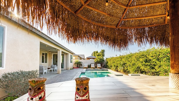 A pool area with two tiki head sculptures, a thatched roof, and a view of a house with outdoor seating. - Luxe Indio Stay w/ Pool, Tiki Bar & Fire Pit Near Coachella | Longfellow (Indio)