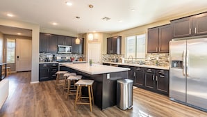 A modern kitchen with dark cabinets, stainless steel appliances, a large island with stools, and a trash can. - Luxe Indio Stay w/ Pool, Tiki Bar & Fire Pit Near Coachella | Longfellow (Indio)