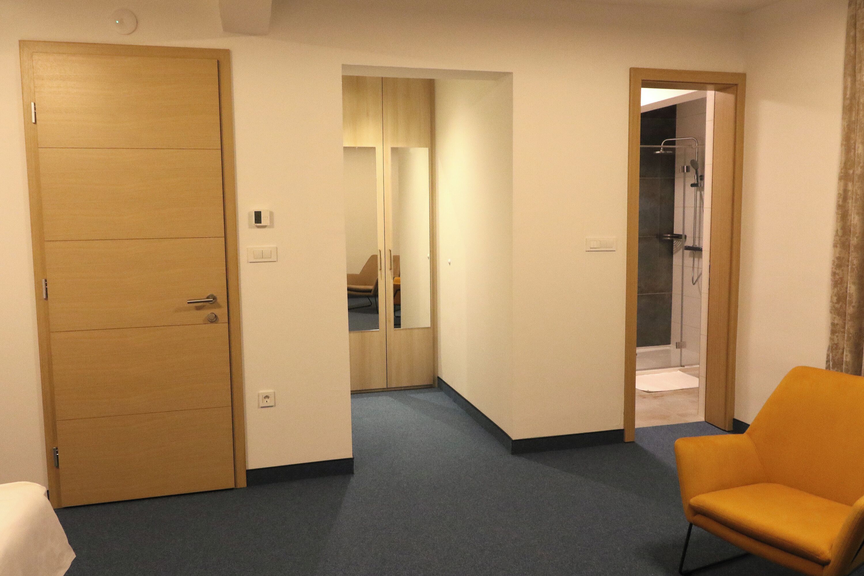 A hotel room with a wooden door, a mirror, a shower area, and a yellow armchair.