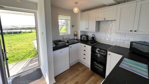 A compact kitchen with white cabinets, a black countertop, and a view of a green landscape through a glass door. - Primrose at Moorhead Country Holidays near Clovelly North Devon (Bideford)