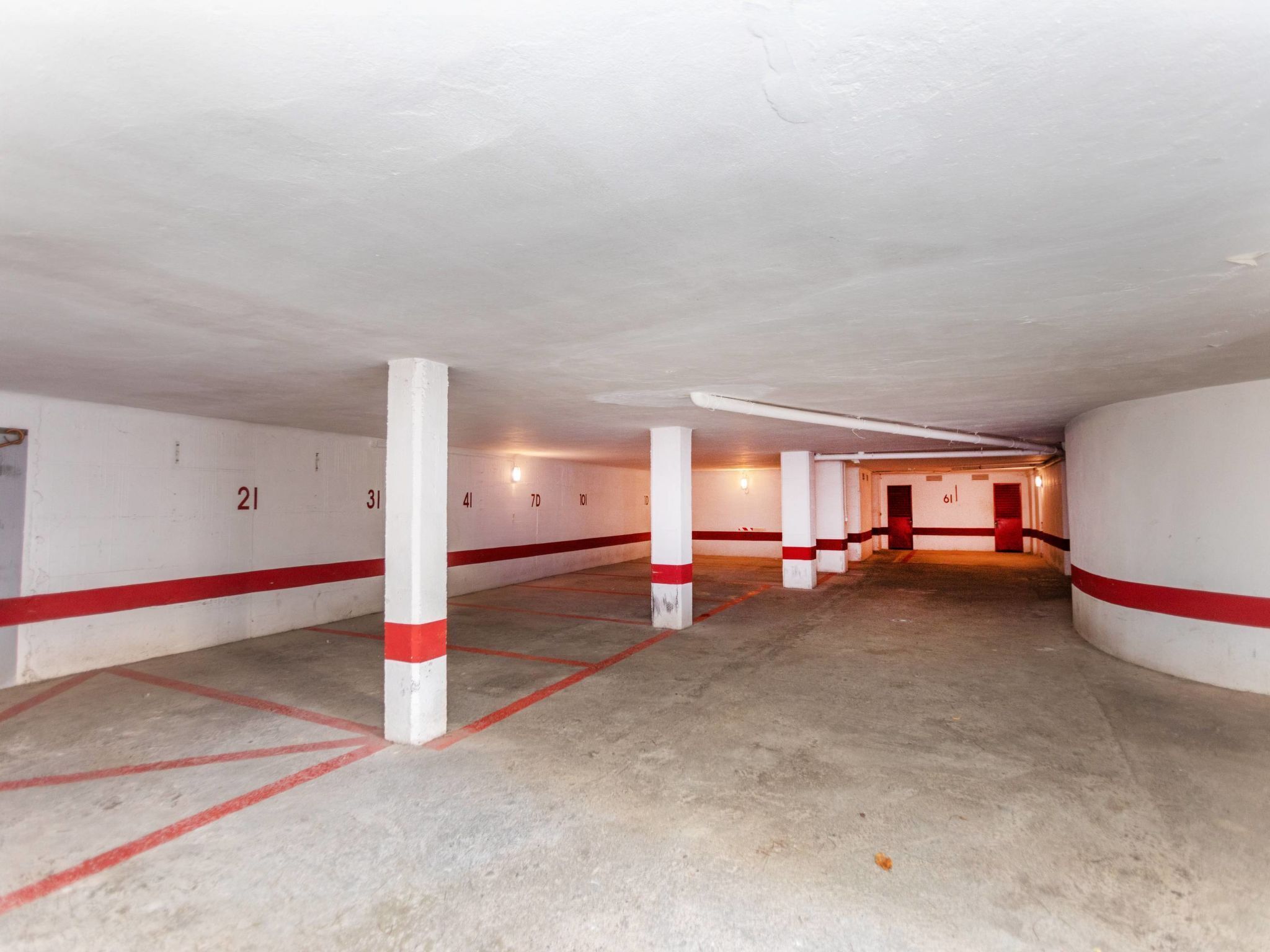 An underground parking garage with white walls and red stripes, featuring numbered parking spaces and concrete pillars.