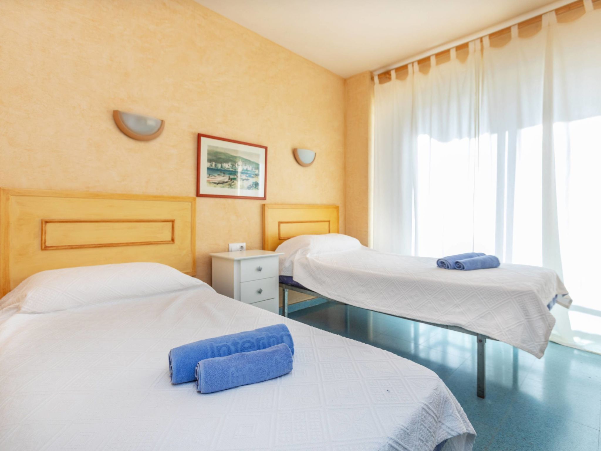 Une chambre d’hôtel avec deux lits, une table de chevet, un tableau encadré au mur et une fenêtre avec des rideaux.