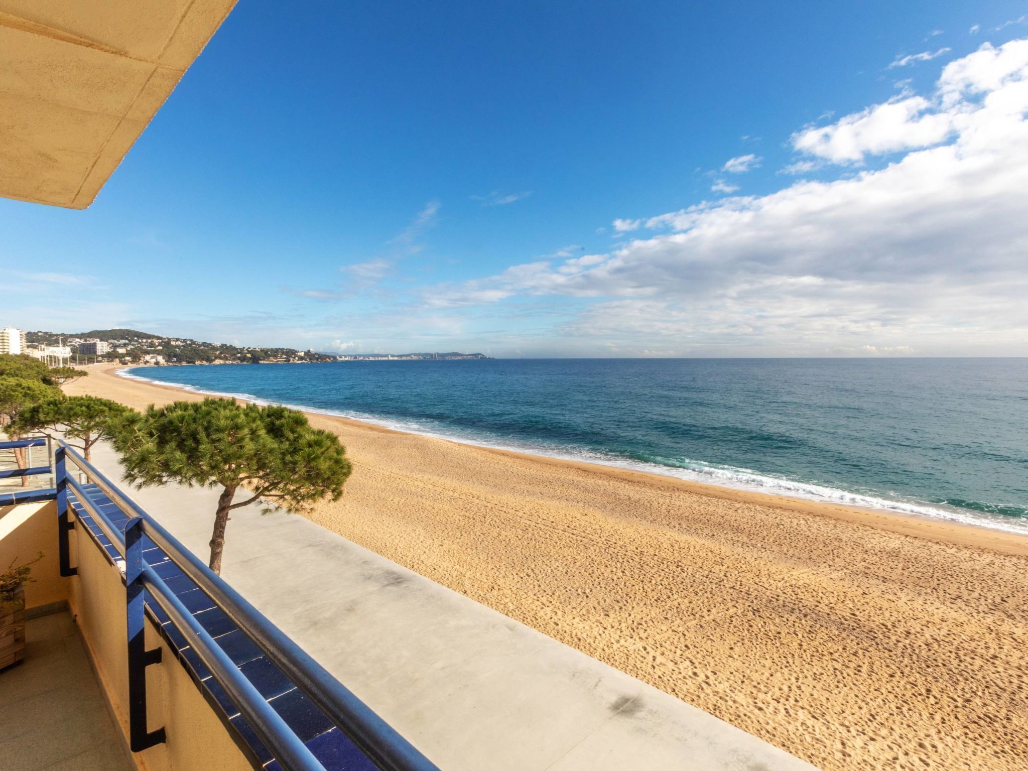 Vista de la playa con balcón que da al océano, playa de arena y edificios a lo lejos.