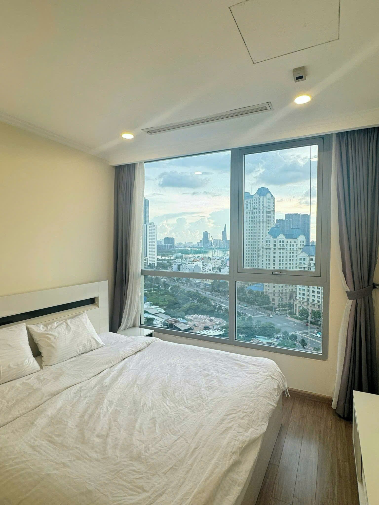 Ein Hotelzimmer mit einem großen Bett, Blick auf eine Stadtkulisse durch ein großes Fenster und einer an der Decke befestigten Klimaanlage.