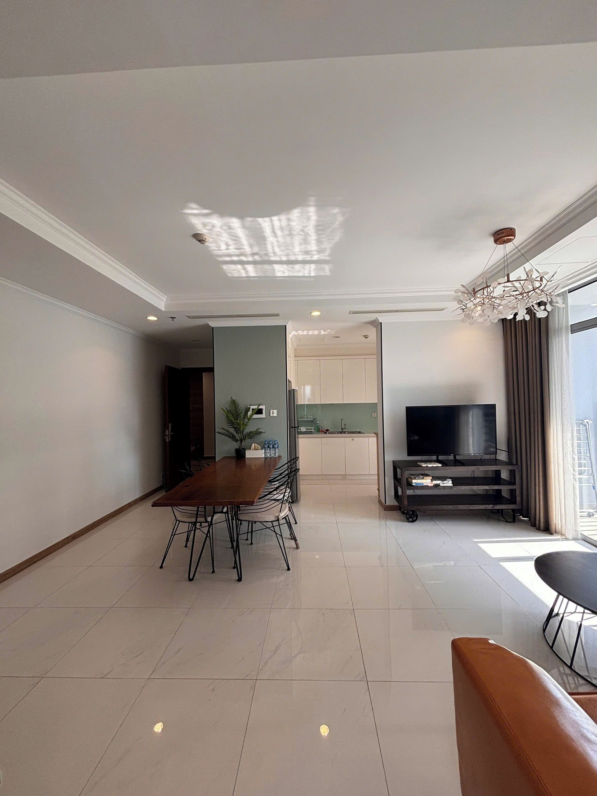 A modern living room with a dining area, a kitchen in the background, and a large window with curtains.