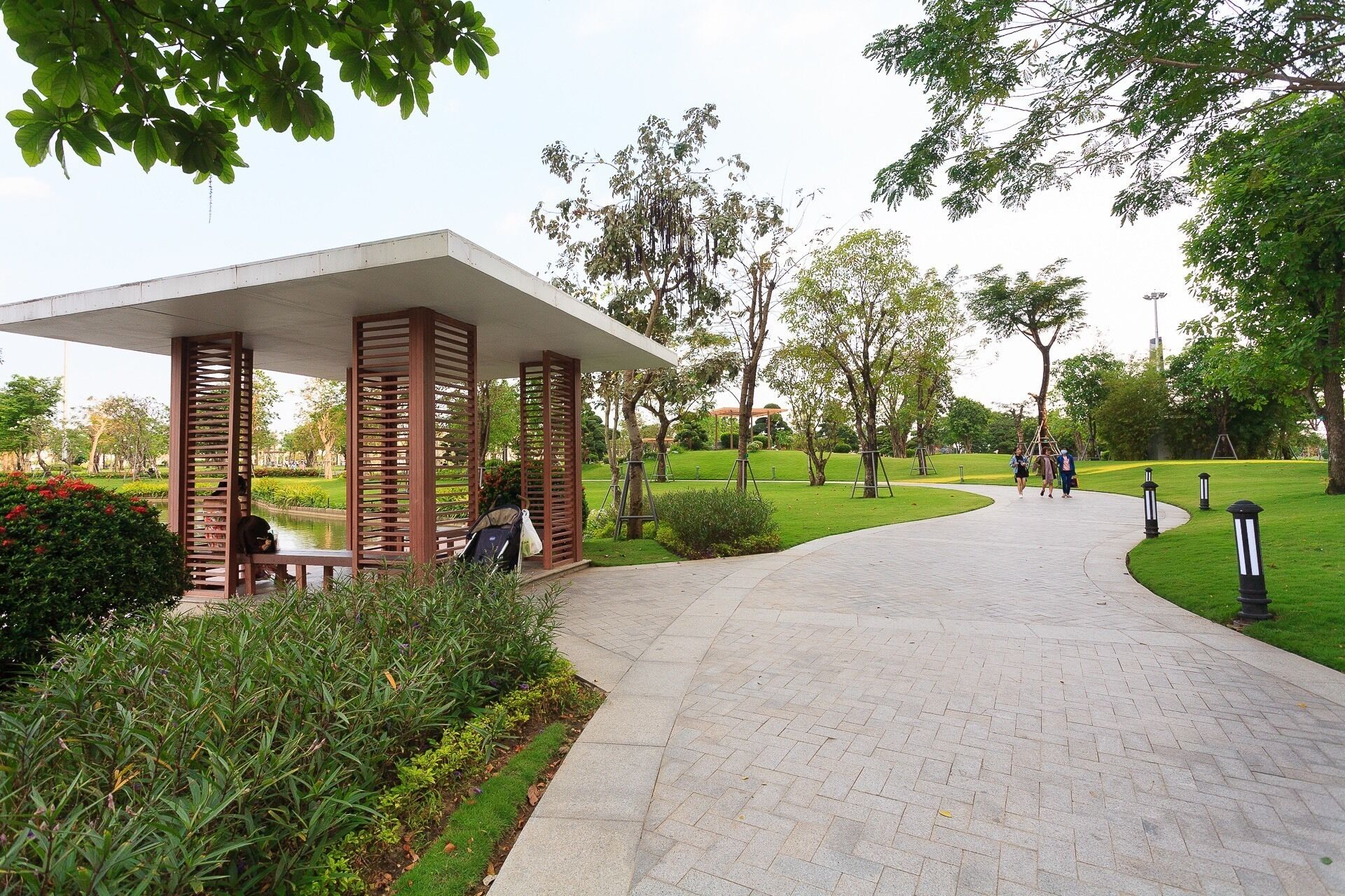 A paved pathway leading to a covered seating area with wooden slats, surrounded by greenery and trees.