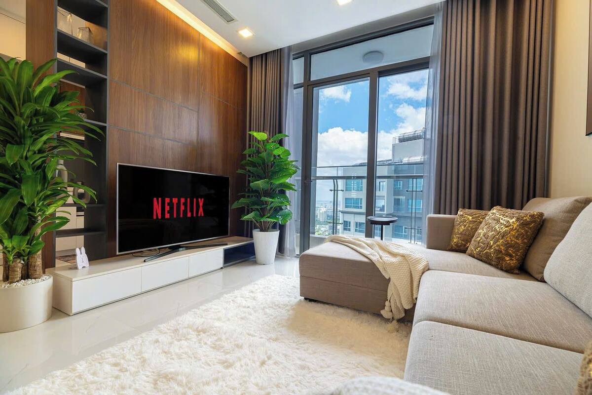 A modern living room with a flat-screen TV, a sofa, a white furry rug, and large windows with a view of buildings.