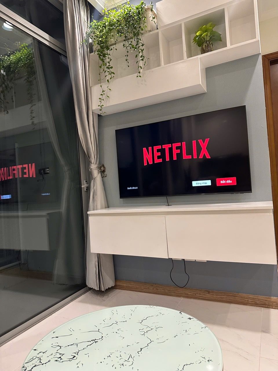 A modern living room with a mounted TV displaying 'NETFLIX' in red letters, a small white table with a marble-like design, and a window with a curtain.