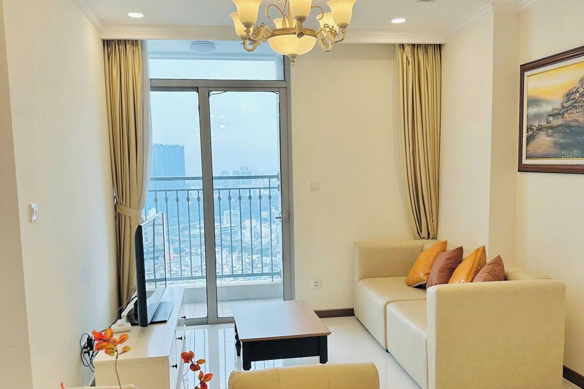 A modern living room with a sofa, a dining table with a vase of flowers, and a balcony with a view.