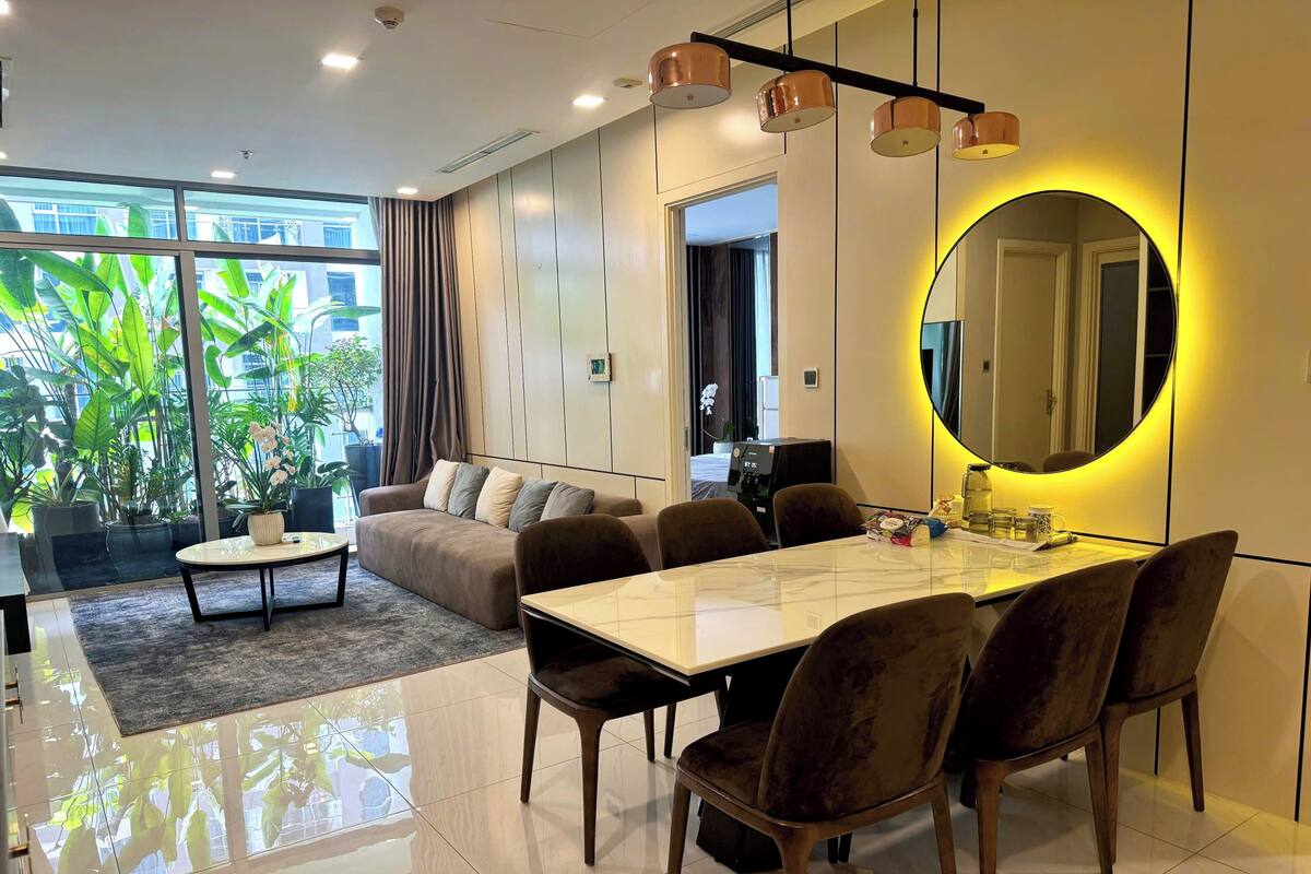 A modern living room with a sofa, coffee table, dining table, and chairs. There is a large window with plants outside.
