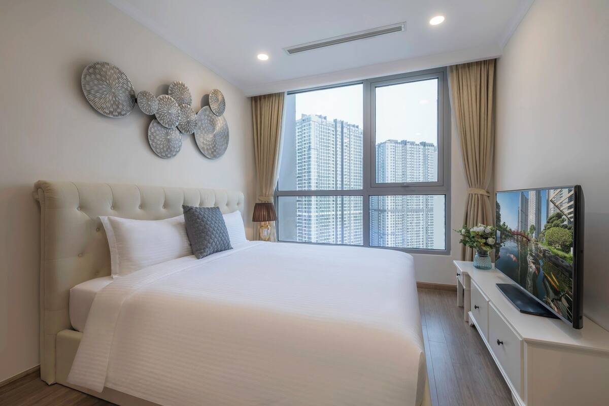 A modern bedroom with a large bed, a television on a white dresser, and a city view through the window.