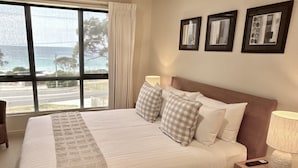 Habitación de hotel con una cama grande, una mesita de noche, una lámpara y un ventanal con vista al mar.