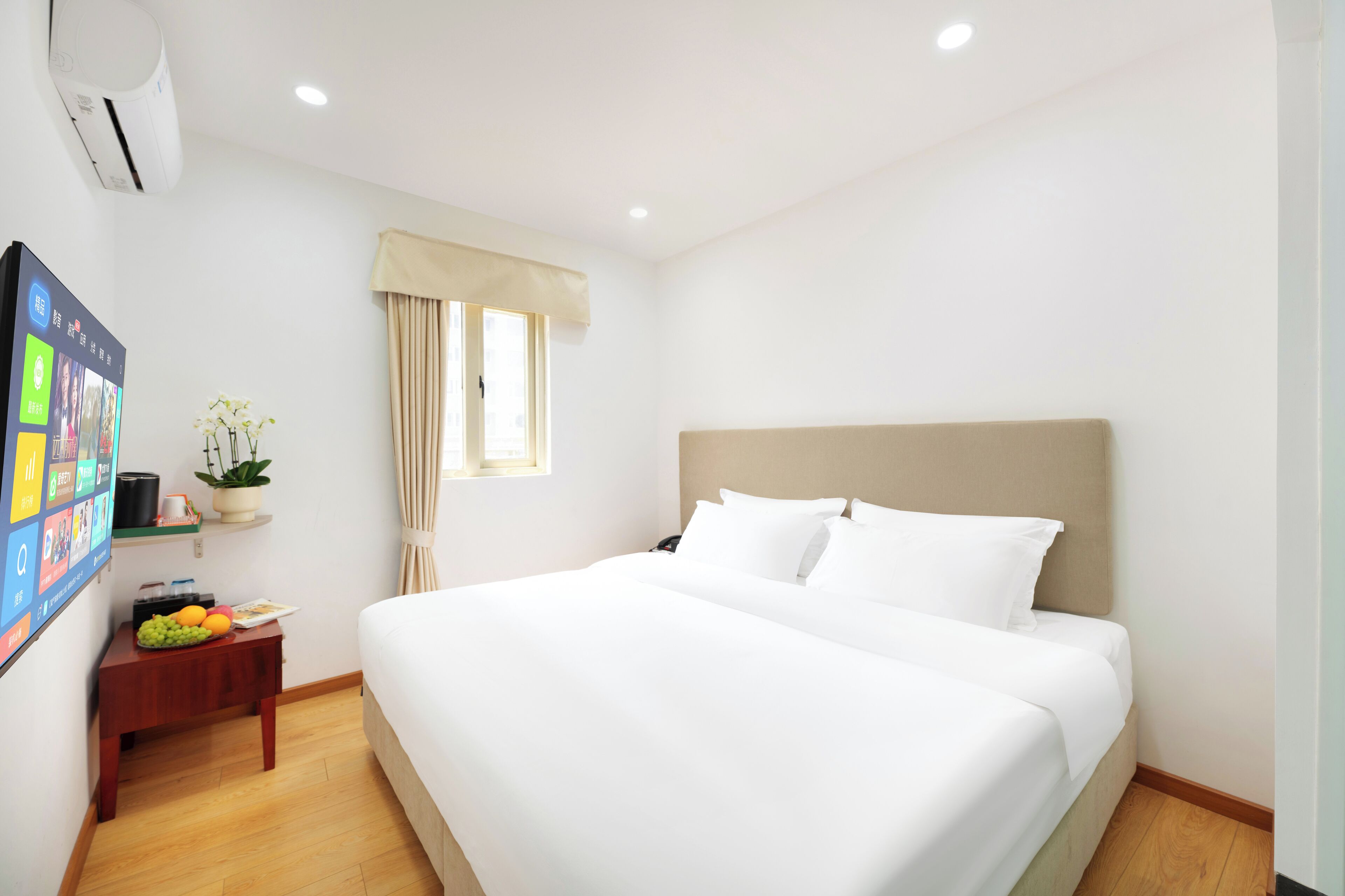 Ein Hotelzimmer mit einem großen Bett, einem Nachttisch mit Früchten, einem Wand-Fernseher und einem Fenster mit Vorhängen.