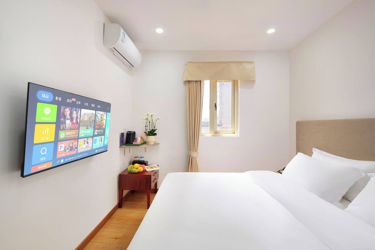 Une chambre d’hôtel avec un lit, un téléviseur fixé au mur, une petite table avec une plante et des fruits, et une fenêtre avec des rideaux.