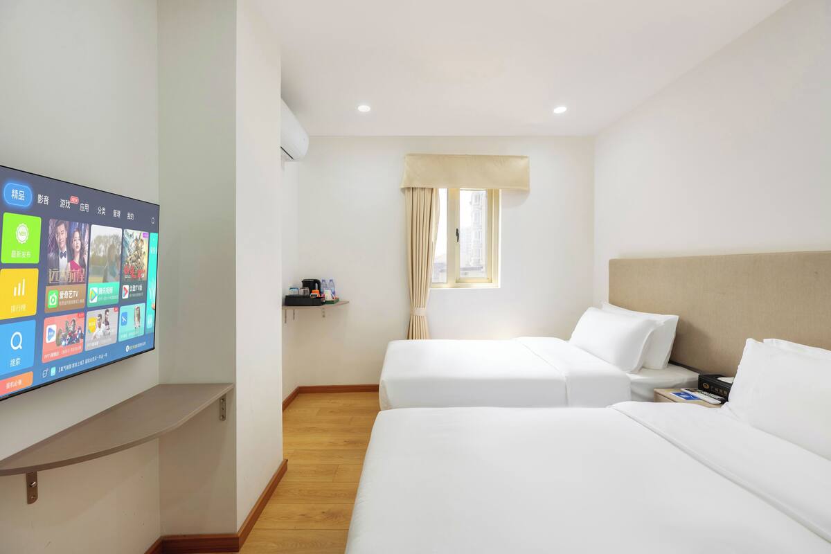 Une chambre d’hôtel avec deux lits, un téléviseur fixé au mur, une fenêtre avec des rideaux et une petite étagère avec des articles de toilette.