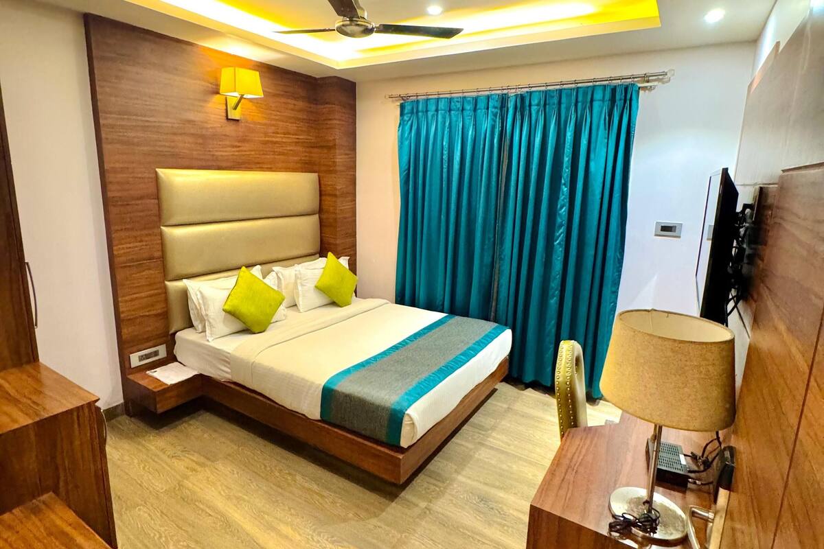 A modern hotel room with a wooden headboard, a bed with white linens and green pillows, a ceiling fan, and a TV on a wooden stand.