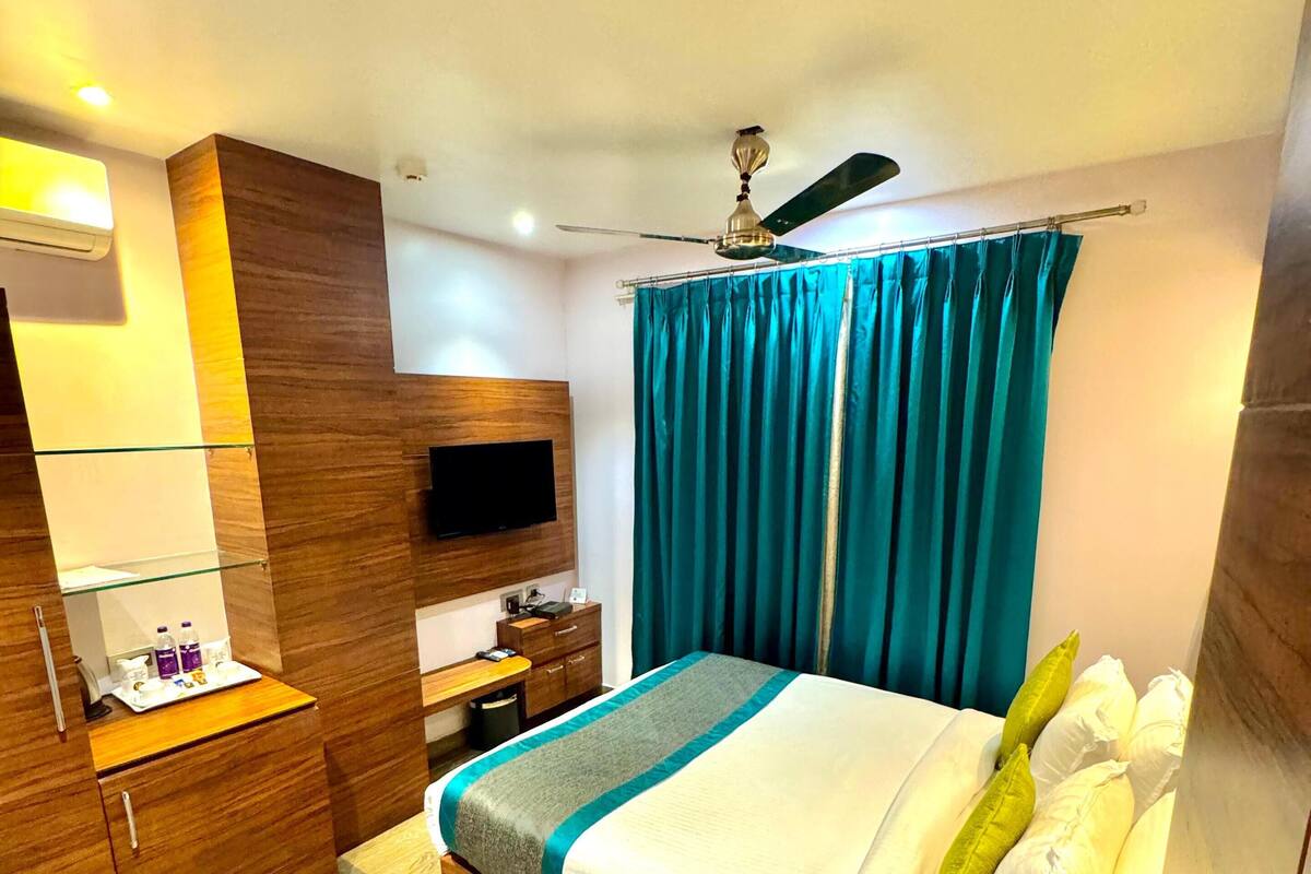 A hotel room with a bed, a desk, a TV, a ceiling fan, and a bathroom with a toilet and sink.
