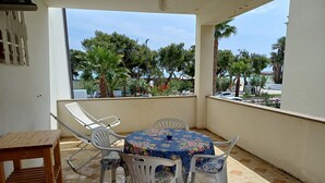 A balcony with a round table and chairs, a wooden side table, and a view of trees and a building in the background. - Apartment 1st row sea with beautiful terrace (Grottammare)