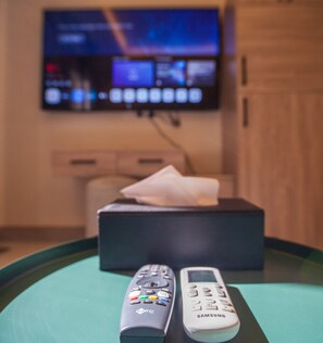 Two remote controls on a table, a tissue box, and a TV in the background. - Elite One-Bedroom Apt AlRimal-3 (Riyadh)