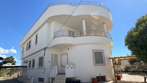 Un edificio de dos pisos con balcón, escalera y un pequeño espacio exterior con plantas en macetas.