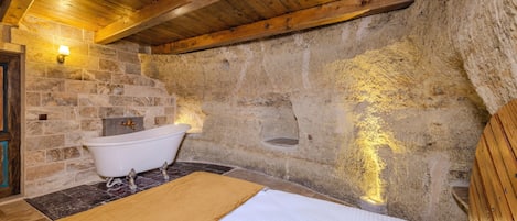 A rustic bathroom with a stone wall, a wooden ceiling, a white bathtub, and a bed with a beige blanket.