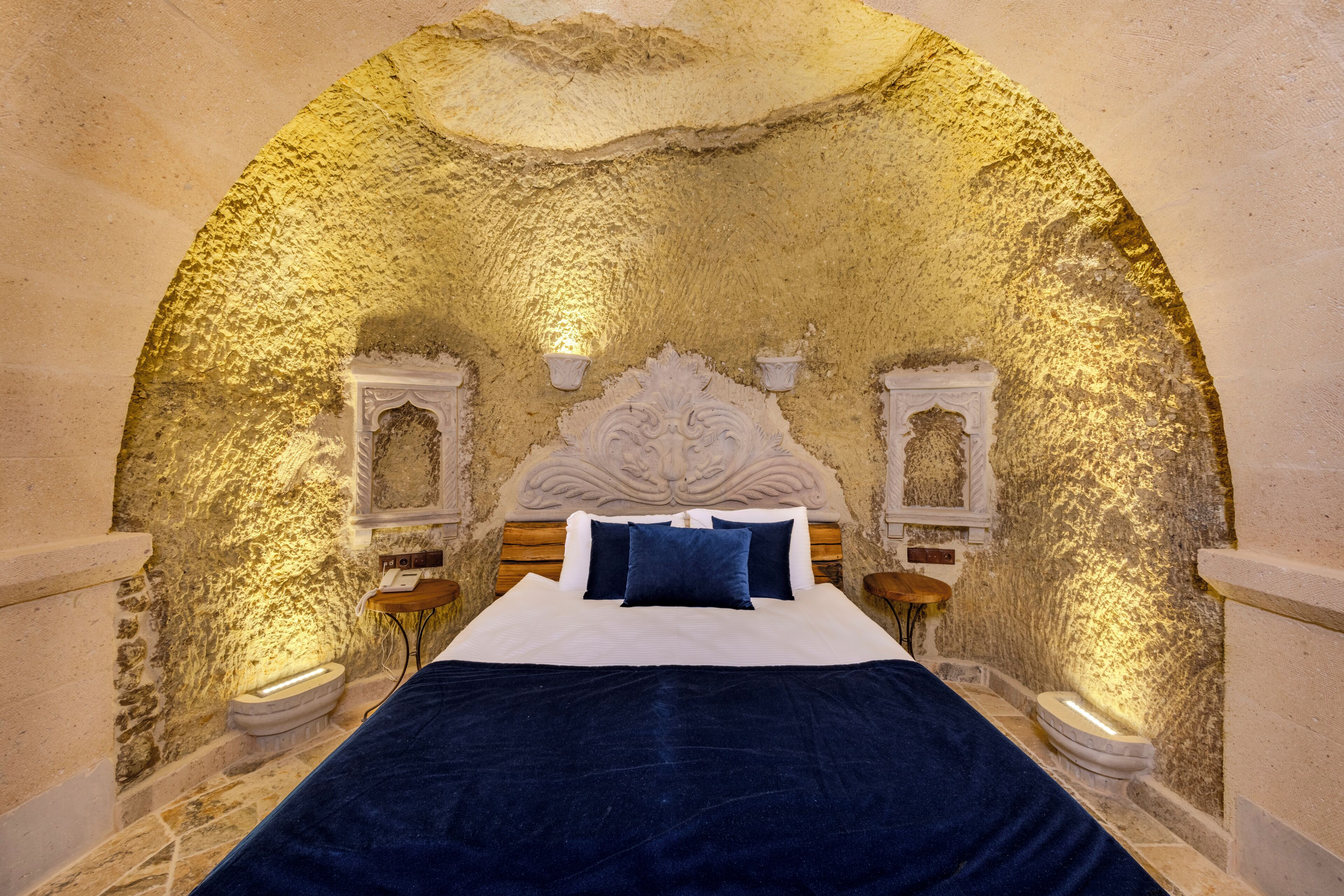 A room with a stone wall, a bed with a white headboard, two bedside tables, and a decorative ceiling.