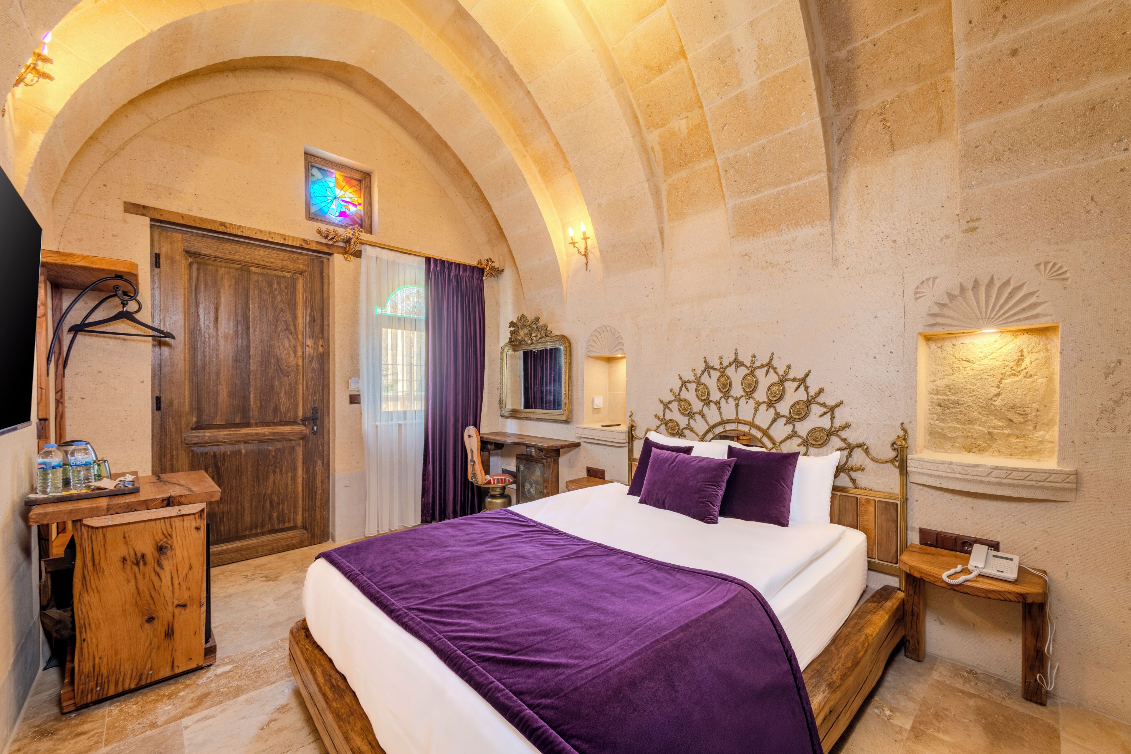 A hotel room with a large bed, a wooden dresser, a vanity with a mirror, and a stone wall.