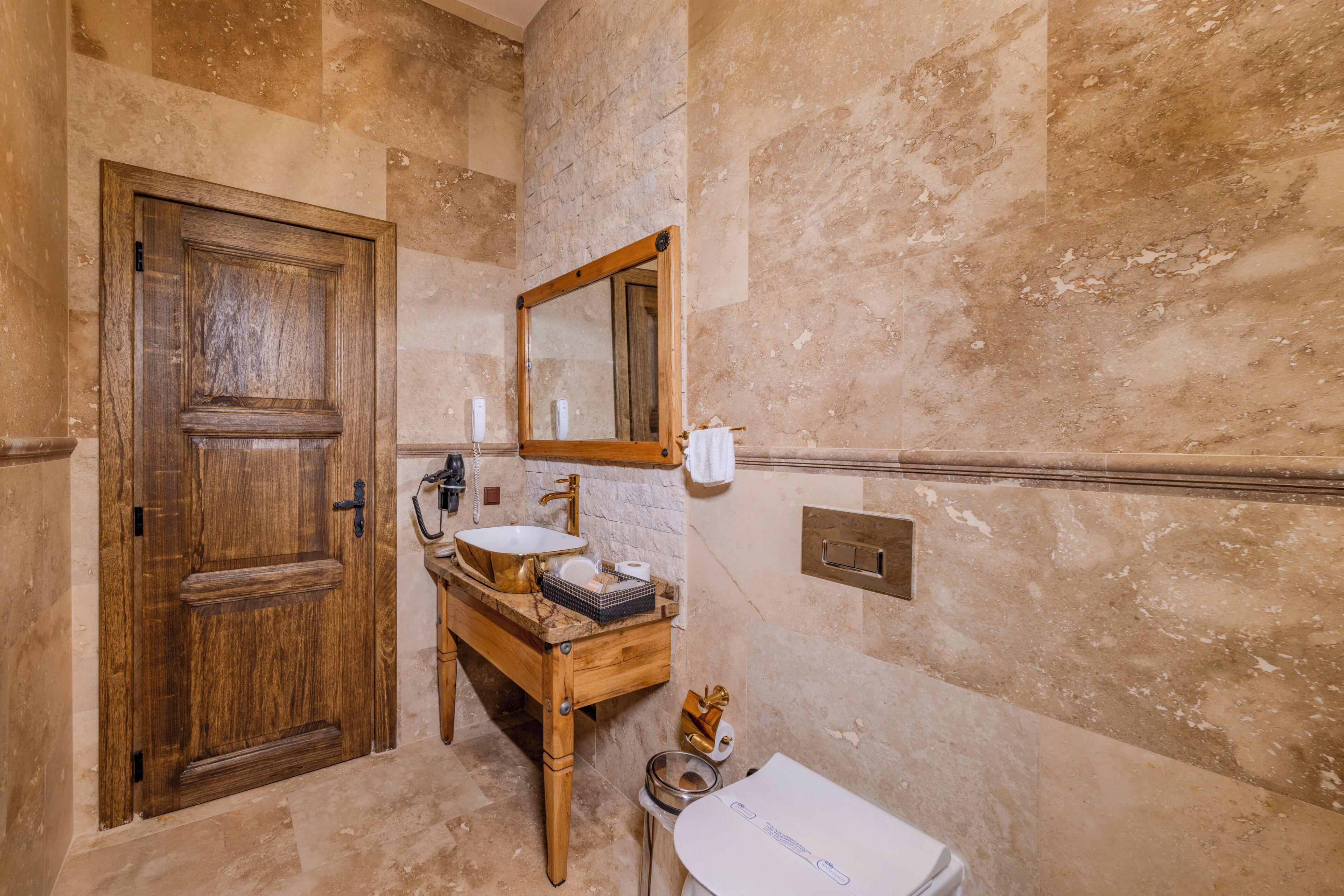 A bathroom with a wooden door, a marble wall, a sink with a gold faucet, and a toilet.