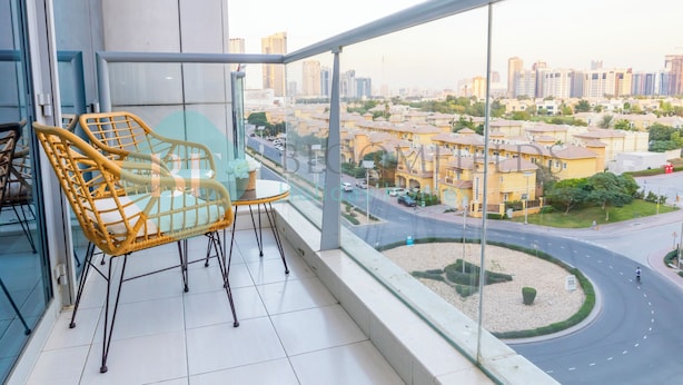 Een balkon met een rieten stoel en een tafeltje, met uitzicht op een stadsgezicht met woongebouwen en een rotonde.