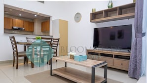 Een moderne woonkamer met een eettafel met glazen blad, houten stoelen, een televisie en een boekenkast.