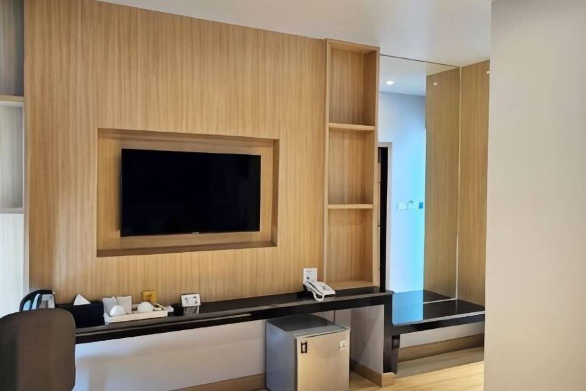 A modern hotel room with a large flat-screen TV mounted on a wooden panel wall, a small desk with a telephone, and a compact refrigerator.